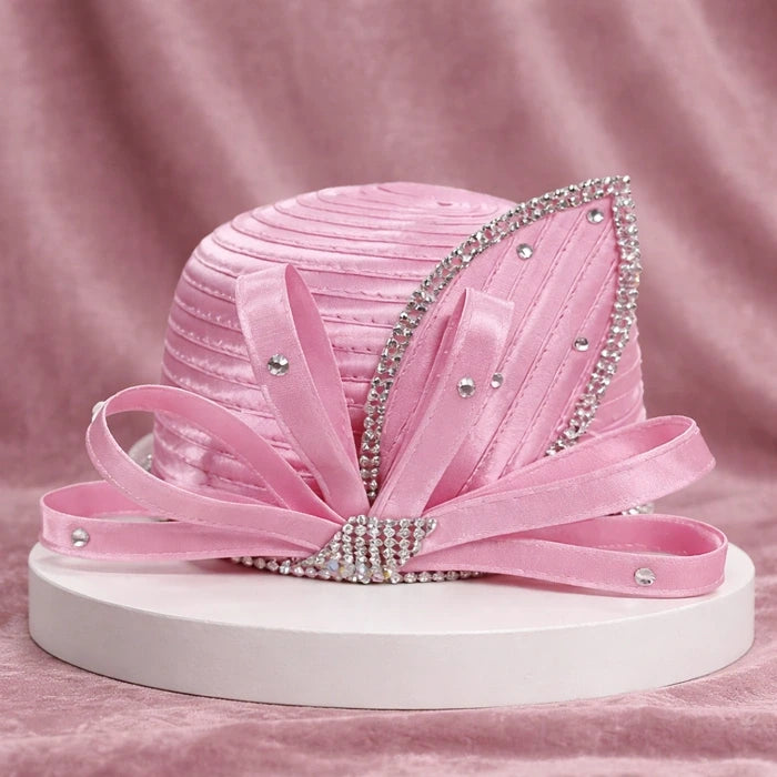Delicate baby pink satin church hat with sparkling rhinestone detailing and flowing satin ribbons, placed on a white pedestal with a soft pink velvet background.