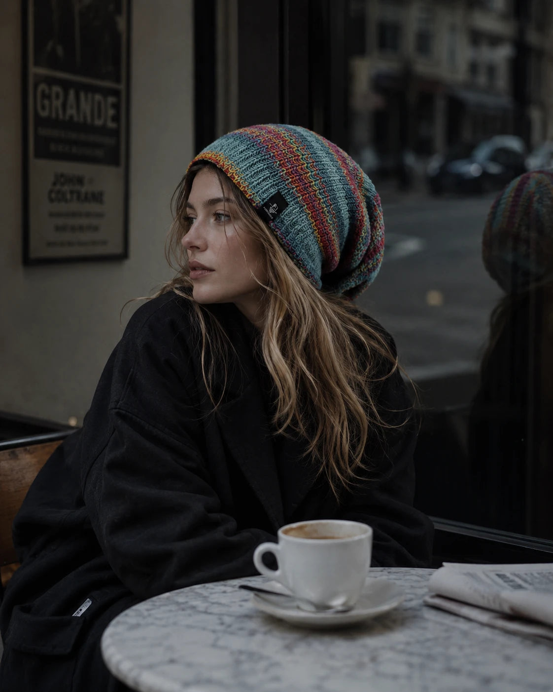 A beautiful woman in a colorful chunky knit slouch hat looking away, with a white marble table, coffee cup, and newspaper in the foreground.