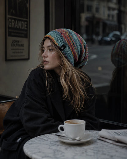 A beautiful woman in a colorful chunky knit slouch hat looking away, with a white marble table, coffee cup, and newspaper in the foreground.
