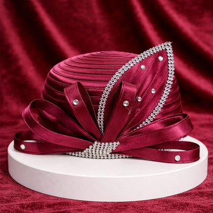 Elegant burgundy satin church hat featuring sparkling rhinestones and layered satin ribbons, showcased on a white pedestal with a rich burgundy velvet background.