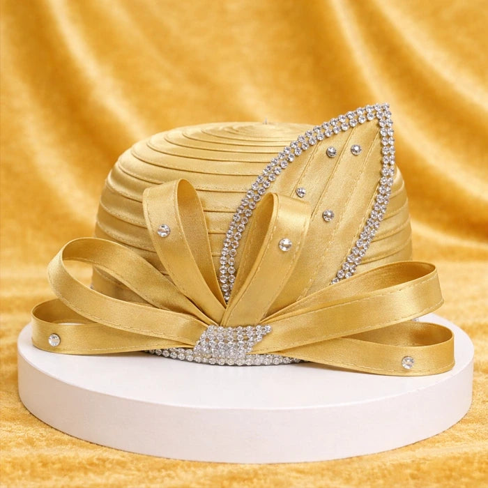 Gorgeous gold satin church hat adorned with rhinestone embellishments and satin ribbons, beautifully displayed on a white platform with a golden velvet backdrop.