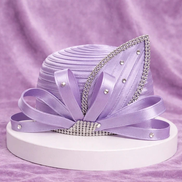 Beautiful lavender satin church hat with sparkling rhinestone accents and elegant satin ribbons, placed on a white pedestal with a lavender velvet backdrop.