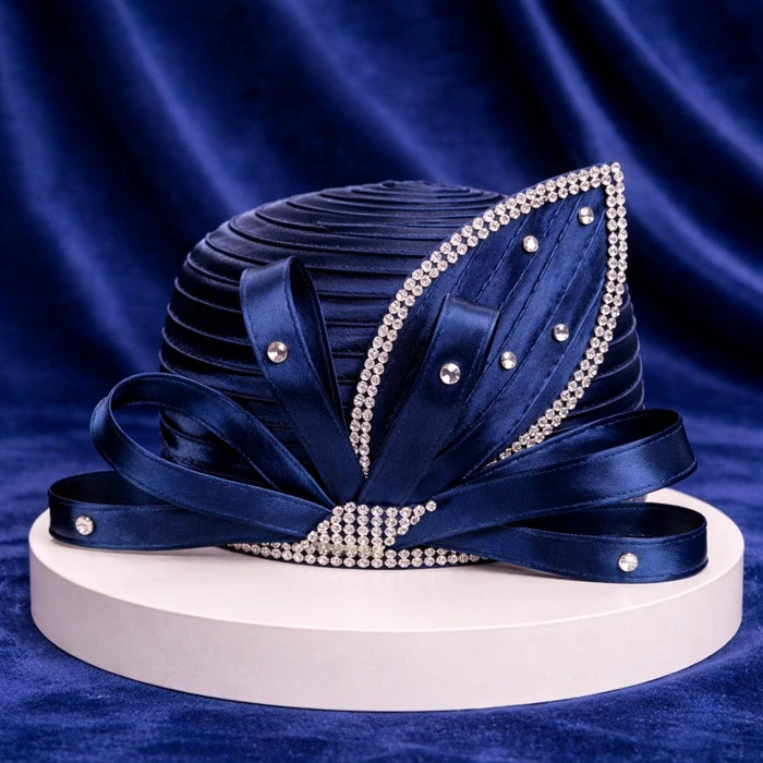 Elegant navy blue satin church hat with intricate rhinestone detailing and satin ribbons, displayed on a white pedestal with a navy velvet background.