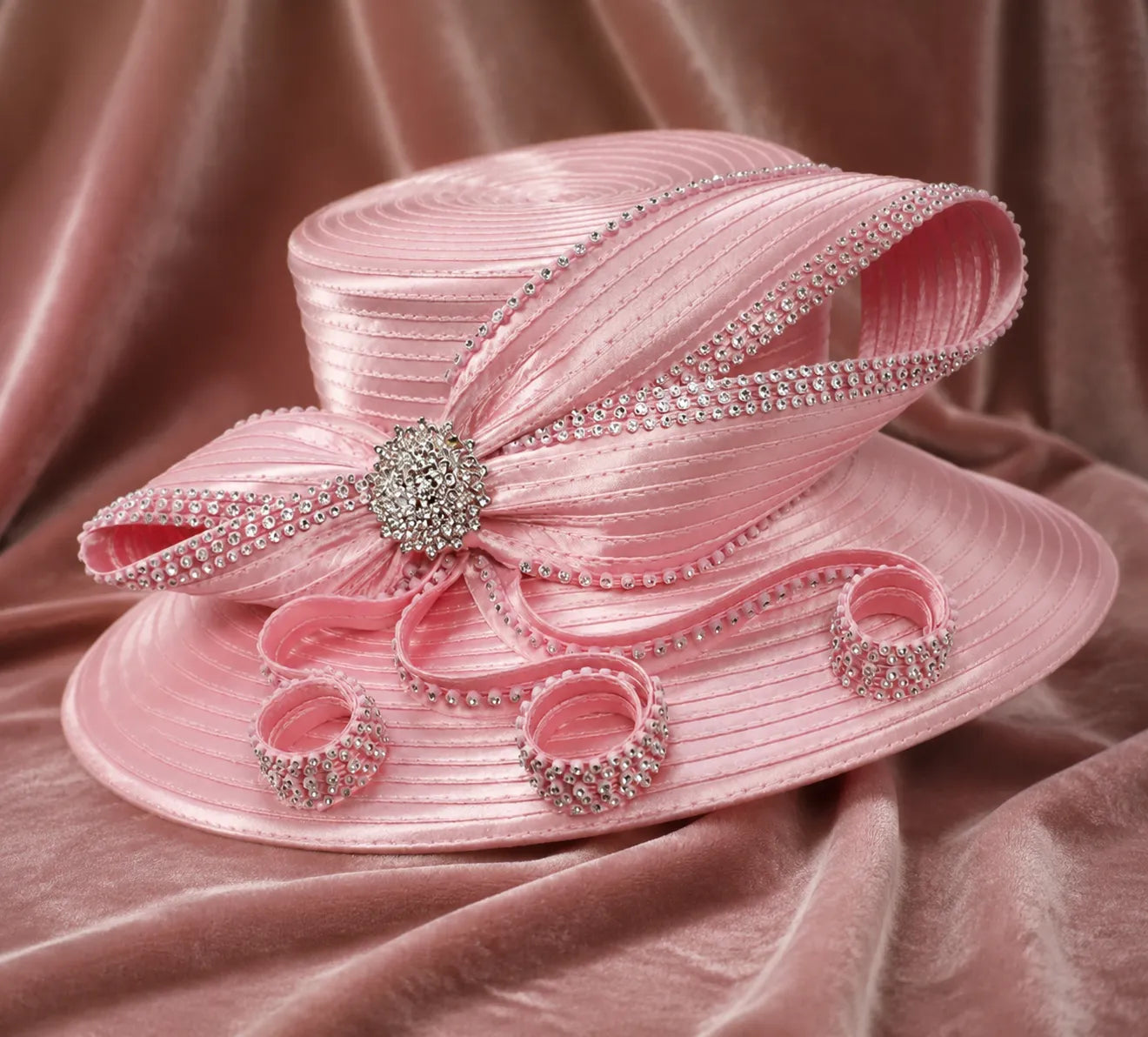 Pink rhinestone church hat with decorative ribbon loops and wide brim displayed on satin fabric background