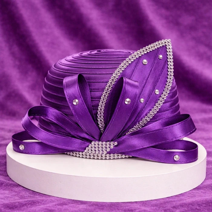 Elegant purple church hat with satin pleats and rhinestone accents, displayed on a white platform with a rich purple velvet background, showcasing its intricate design.