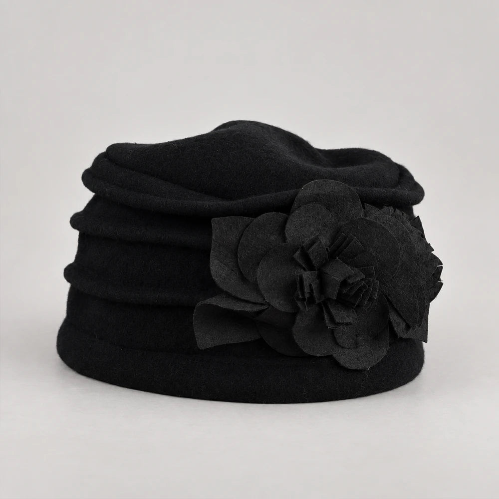 A black felt hat featuring a large, layered fabric flower applique on the side, photographed against a neutral light gray background.