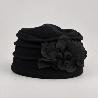A black felt hat featuring a large, layered fabric flower applique on the side, photographed against a neutral light gray background.